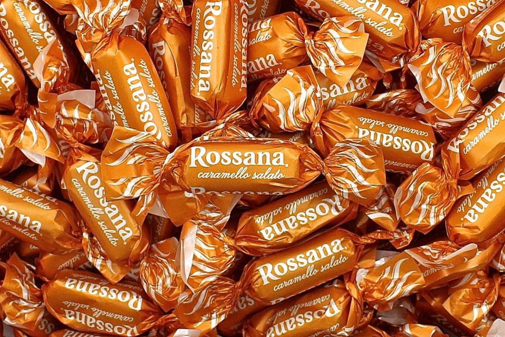 Primo piano di numerose Caramelle Rossana Caramello Salato sfuse nel loro incarto color caramello con scritta e logo in bianco.