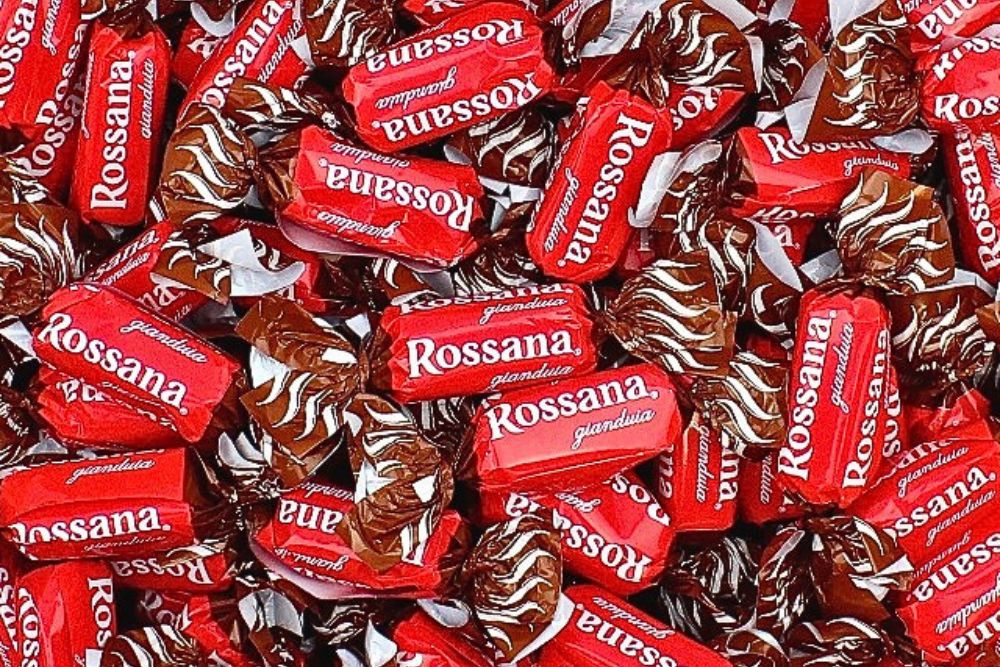 Primo piano di numerose Caramelle Rossana Gianduia sfuse nel loro incarto rosso con con scritta e logo in bianco