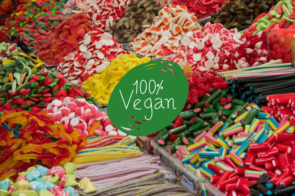 Un assortimento colorato di caramelle gommose vegane esposte in pile con un'etichetta verde al centro che recita "100% Vegan"
