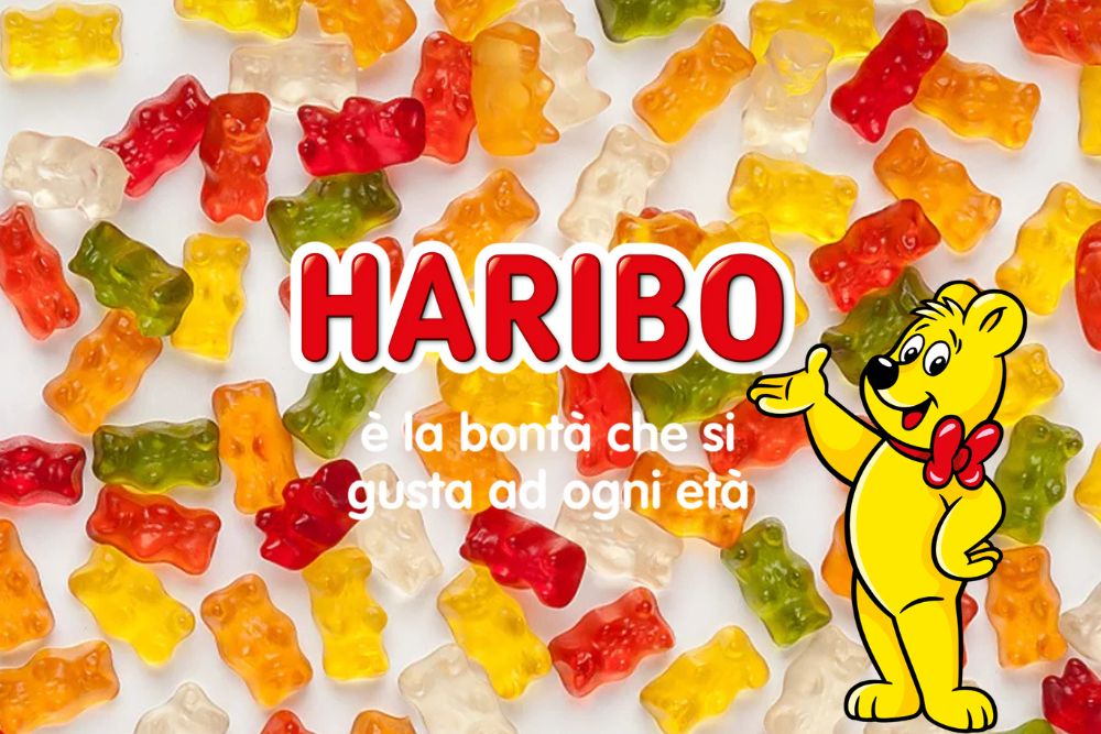 Orsetti gommosi Haribo Orsetti d’Oro sparsi su uno sfondo bianco con il logo HARIBO, una simpatica mascotte orsetto giallo con papillon rosso e lo slogan "Haribo è la Bontà che si gusta ad ogni Età".