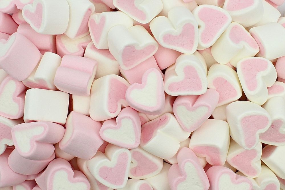 Marshmallow rosa e bianco a forma di cuore, disposti alla rinfusa su uno sfondo neutro.