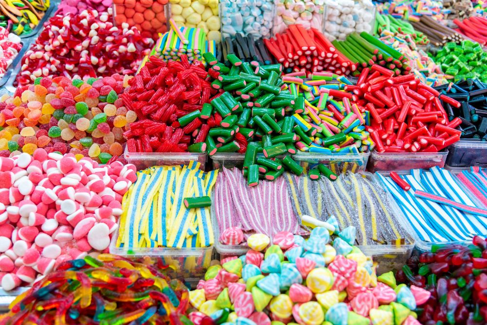 Varie caramelle colorate assortite disposte in contenitori trasparenti, con forme e consistenze diverse, in un mercato dolciario