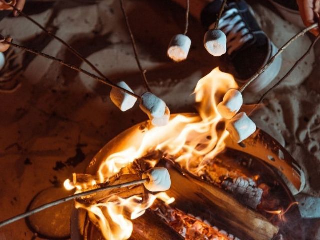 Marshmallow arrostiti su bastoncini sopra un falò acceso sulla sabbia di sera.