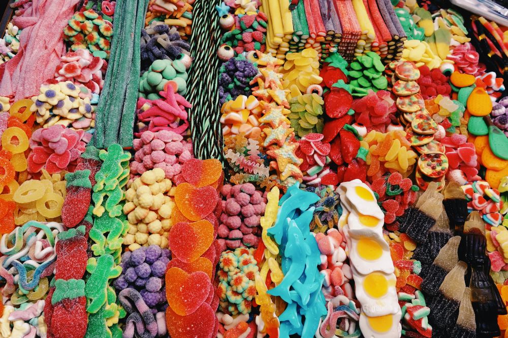 Una bancarella di mercato con un  vivace assortimento di caramelle gommose, liquirizie, strisce aspre, caramelle a forma di frutta e marshmallow ricco di colori vibranti e formati golosi che creano un'atmosfera golosa e allegra.