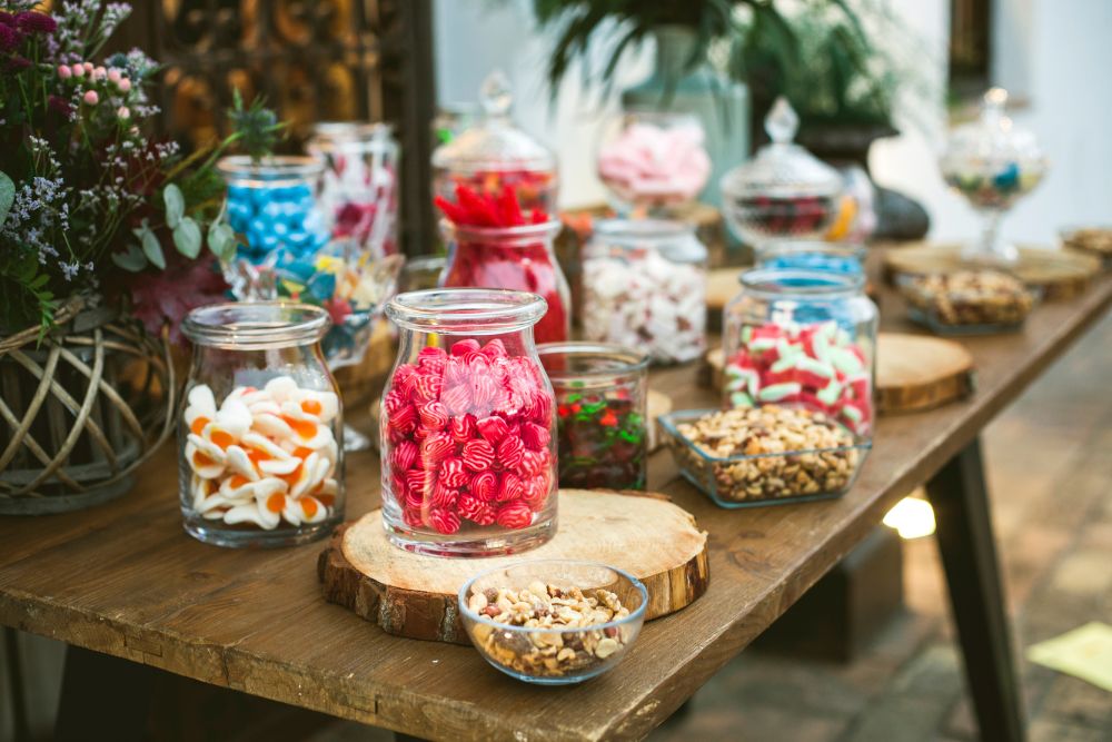 Un buffet di caramelle all’aperto con barattoli di vetro, alzatine e contenitori trasparenti, pieni di caramelle sfuse di vari tipi – gommose, lecca lecca, marshmallow, etc - e altri snack dolci e salati, allineati su un tavolo di legno rustico, creando un allestimento allegro e invitante.