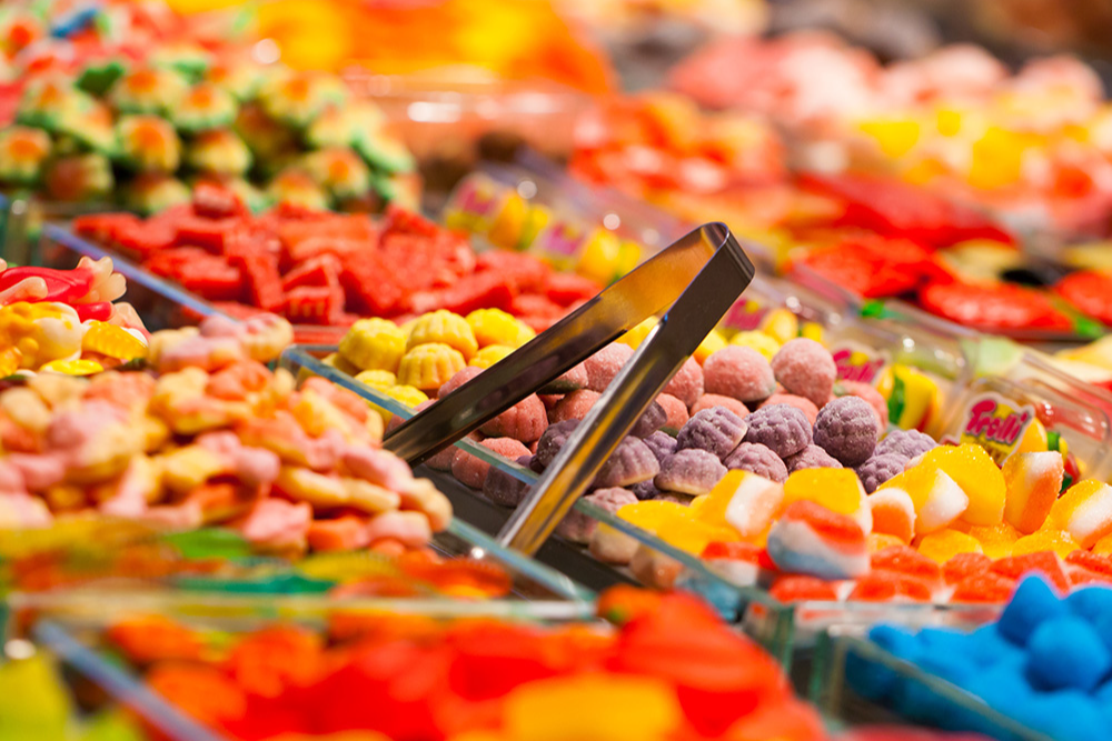 Un colorato allestimento di dolciumi sfusi, con pinze di metallo appoggiate tra eleganti contenitori trasparenti in vetro pieni di caramelle gommose di varie tipologie, forme e colori per creare una scena vivace e invitante.