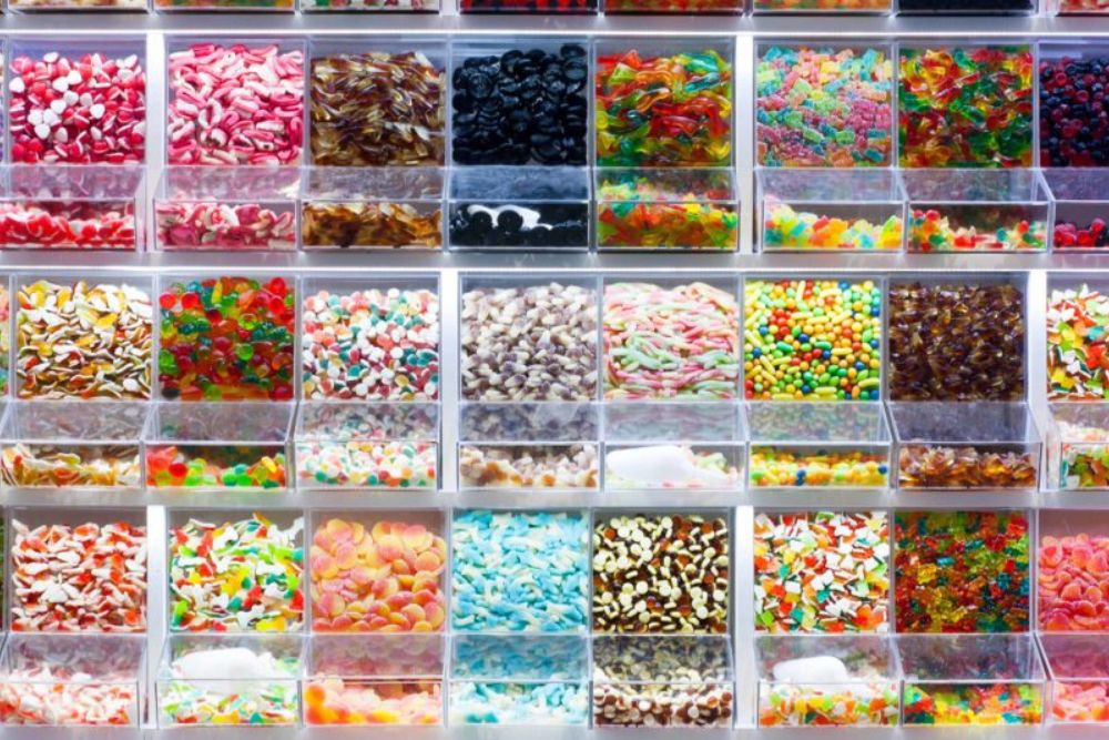 Un vero e proprio muro di caramelle con file ordinate di contenitori trasparenti che espongono un assortimento di caramelle gommose e dolciumi sfusi creando una parete del negozio ordinata e vivace, dall'aspetto invitante.