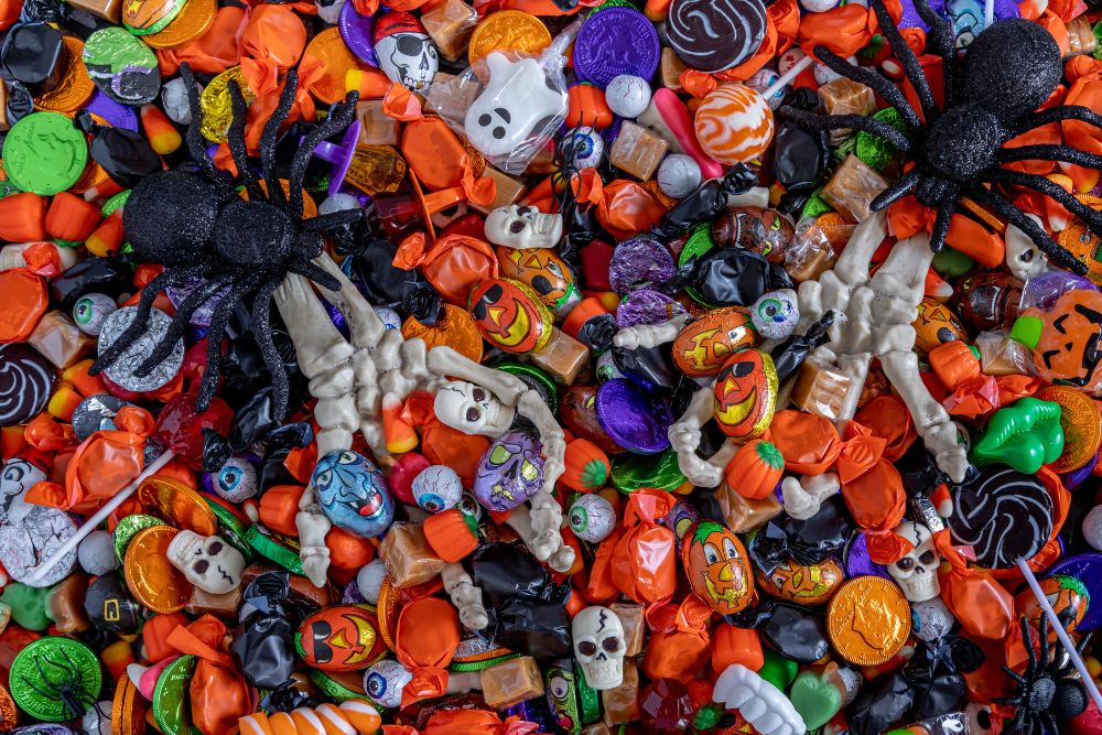 Foto dall'alto di una varietà di caramelle di Halloween colorate con zucche, teschi, ragni neri e dolcetti avvolti in carta arancione, viola e verde a tema.
