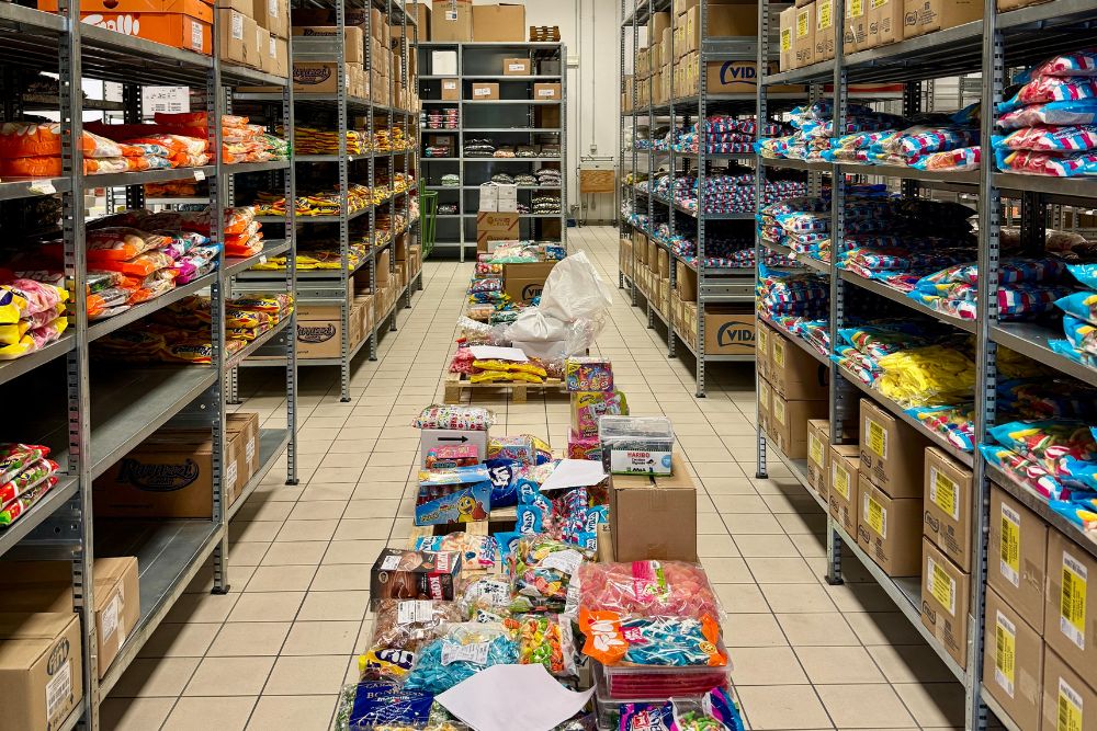 Corridoio del magazzino di Prodotti Dolciari Rigato , fiancheggiato da scaffali pieni di caramelle e snack in confezioni sfuse e scatole bulk, con merci assortite impilate su pallet pronte per essere spedite ai clienti dell’ingrosso