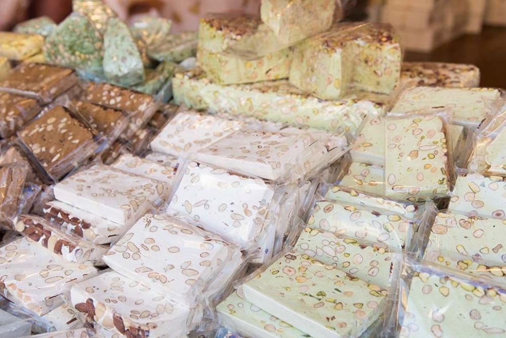 Varie tipologie di torrone bianco, al cioccolato e pistacchio con mandorle, nocciole e frutta secca, esposte su un banco.