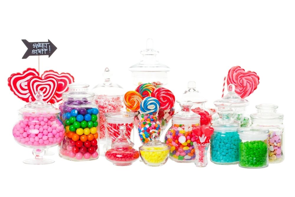 Esposizione di caramelle colorate su sfondo bianco: barattoli di vetro di varie forme e altezze che  contengono caramelle gommose, marshmallow, lecca-lecca e dolciumi nei colori rosa acceso, rosso, blu, giallo e verde.