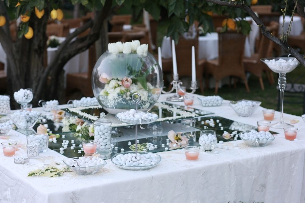 Elegante confettata all'aperto con confetti bianchi, un centrotavola floreale, contenitori in vetro, candele e alberelli di agrumi che creano un'atmosfera romantica e naturale.