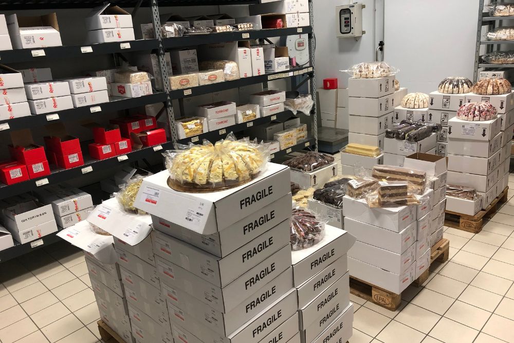 Interno magazzino a temperatura controllata Rigato con scaffali pieni di varie tipologie di torrone incartato sia di produzione propria che di marchi nazionali, pronte per la vendita all’ingrosso in scatole bene organizzate.