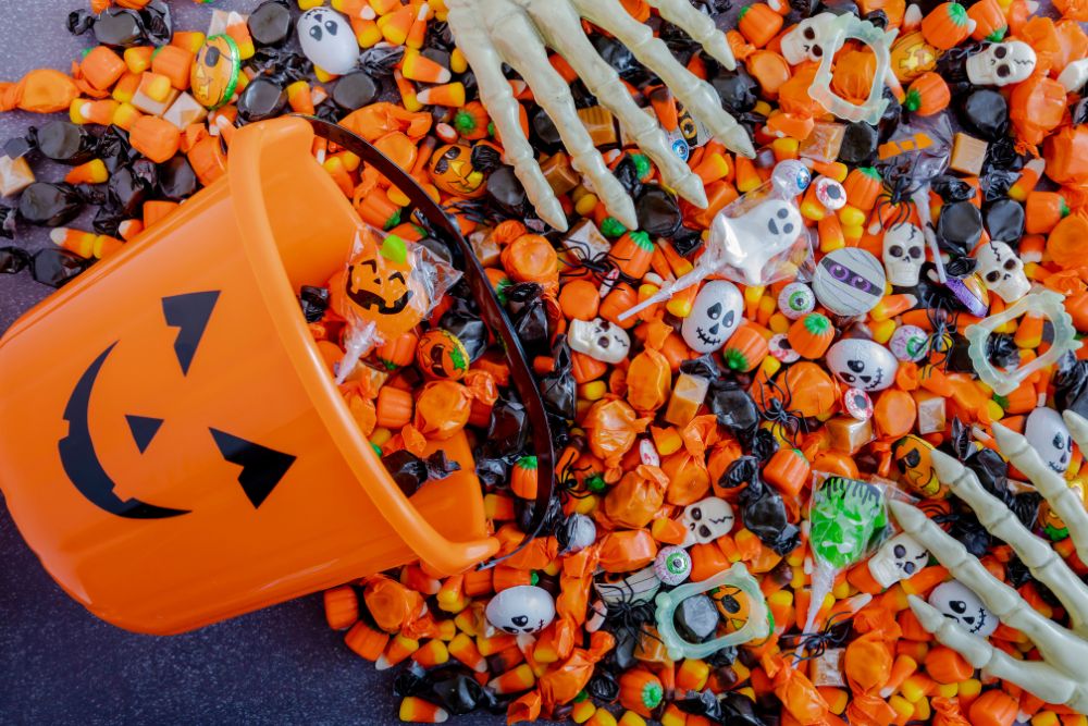 Un secchiello arancione di plastica a forma di zucca di Halloween rovesciato con tantissime caramelle colorate a tema e due mani di scheletro attorno che creando una scena golosa a tema halloween.