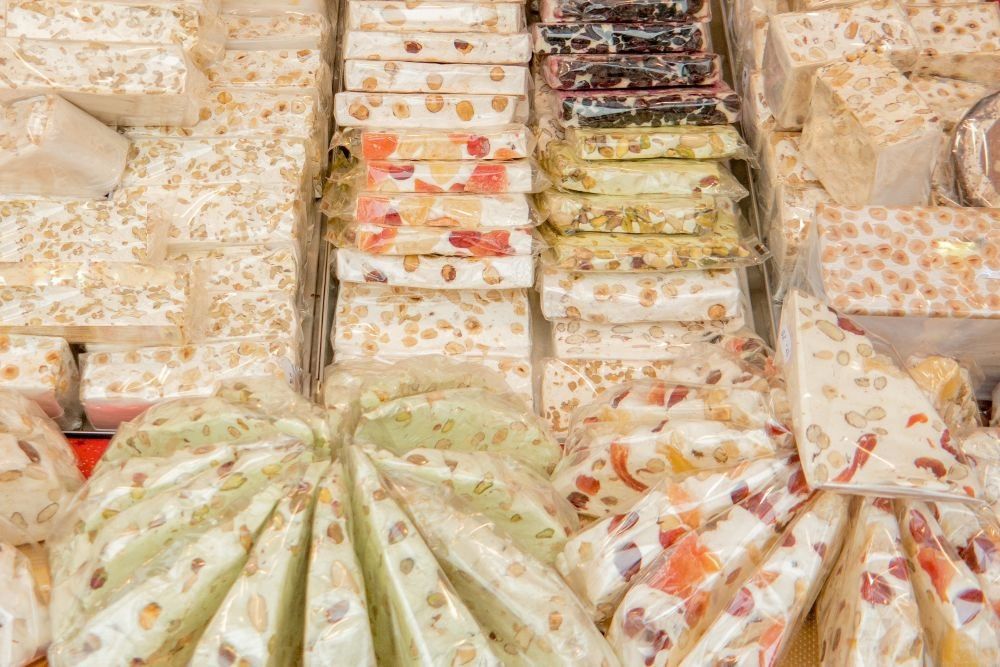 Varie tipologie di torrone artigianale (torte torrone morbido, pani di torrone friabile da taglio, stecche di torrone) con frutta secca incartati, disposti ordinatamente in vassoi da esposizione