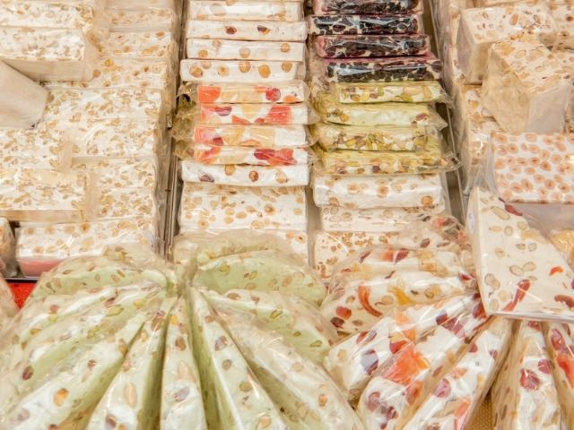 Varie tipologie di torrone artigianale (torte torrone morbido, pani di torrone friabile da taglio, stecche di torrone) con frutta secca incartati, disposti ordinatamente in vassoi da esposizione