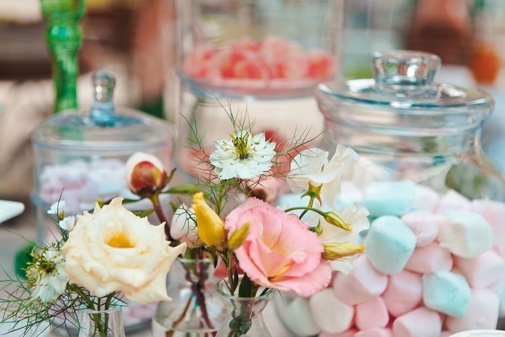 Primo piano stretto di un dettaglio sweet table: Una composizione floreale dai toni pastello, con rose e fiori di campo, affiancati da barattoli di vetro contenenti marshmallow rosa e blu, che creano una composizione still life raffinata e elegante.