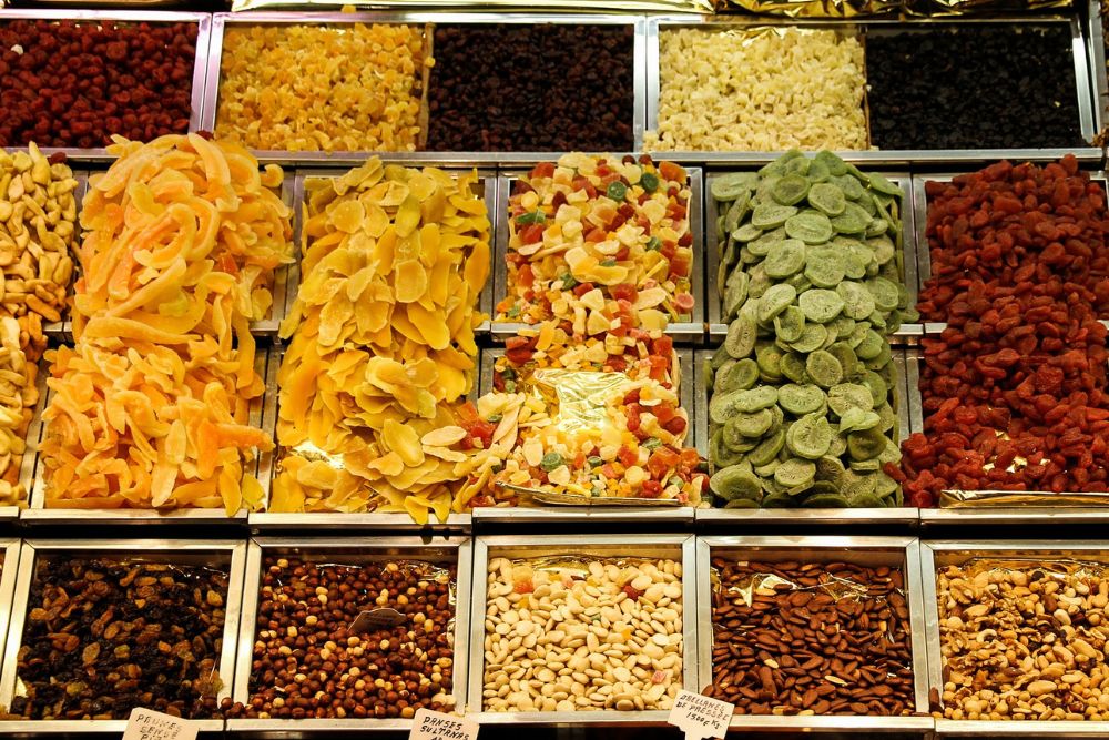 Un colorato banco al mercato che espone frutta secca di varie tipologie come: noci, nocciole, mandorle; semi disposti in contenitori aperti e frutta disidratata tra cui: albicocche, ananas, mango, uvetta per un’offerta ricca e invitante.