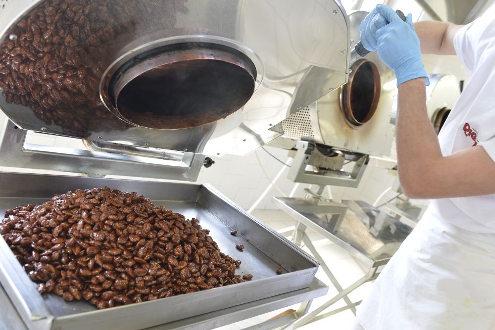 Un addetto Rigato con una grande bassina mentre prepara mandorle pralinate che vengono versate su un vassoio di metallo per il raffreddamento; un’immagine che coglie una produzione artigianale che richiede concentrazione, perizia e materie prime di qualità.