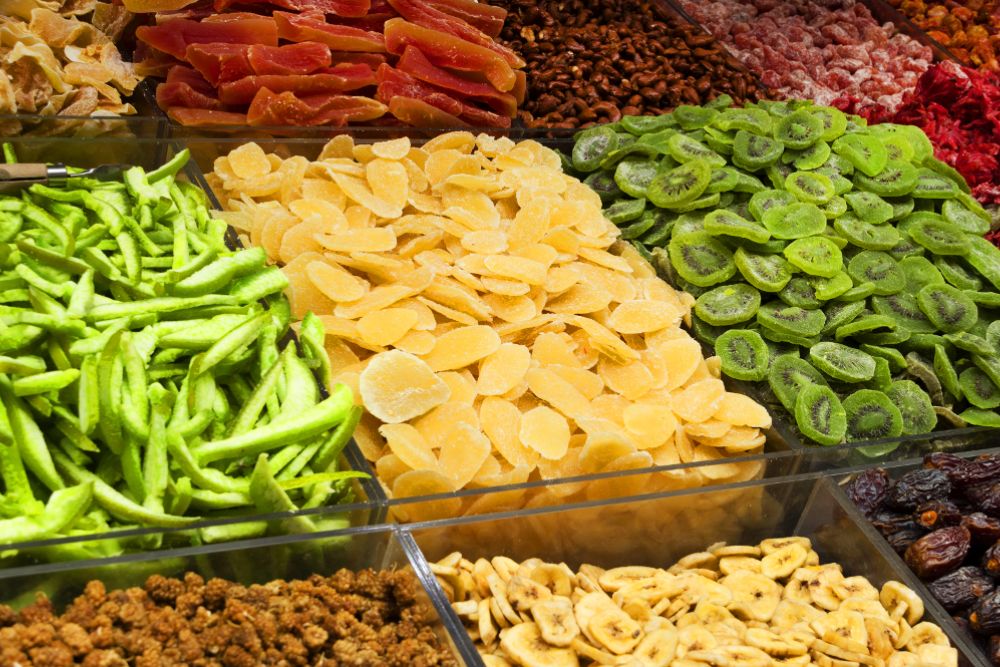 Un colorato banco del mercato con frutta secca e disidratata: fette di mango dorate, rondelle di kiwi e di banana, prugne e datteri essiccati, mango e papaya a fette e un misto di frutti di bosco disidratati, disposti in contenitori aperti e trasparenti.